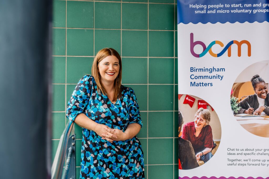 Becky Coley is wearing a blue and green dress, smiling, and standing next to a BCM pull-up banner